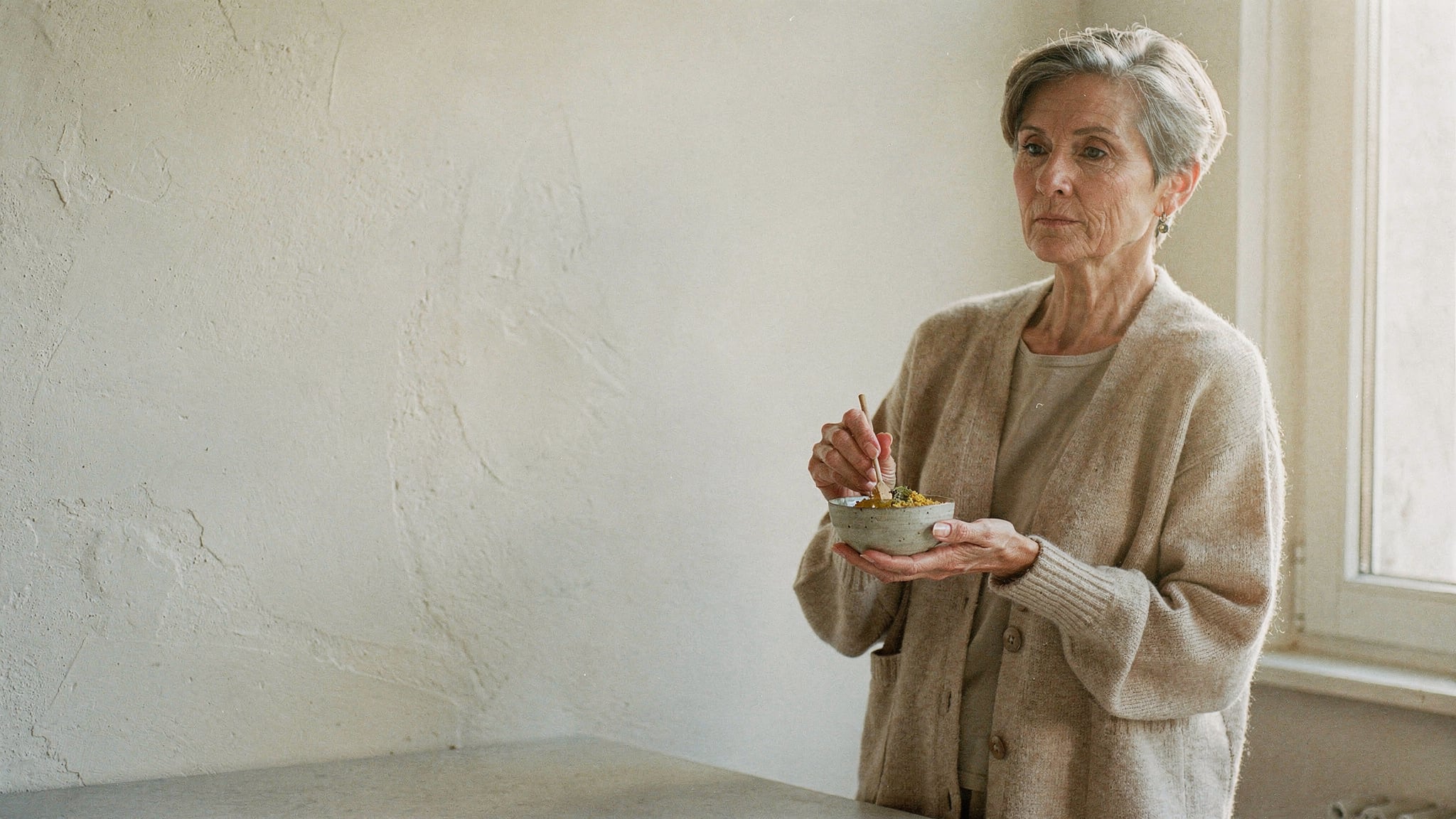 A woman in her sixties stands at her kitchen counter in morning light, holding a small ceramic bowl of homemade honey and mint rub and gazing calmly toward the window.